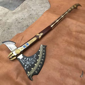 May include: A long-handled battle axe featuring a black blade with white runic patterns and gold trim. The handle is brown with gold metal accents, including circular emblems and blue circular inlays, ending in a curved, pointed gold pommel.