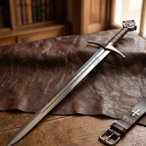 May include: A steel sword with a double-edged blade featuring engraved Latin text, a brown leather-wrapped grip, and a circular pommel with a red cross emblem. A matching brown leather belt with a metal buckle and cross accents lies nearby.