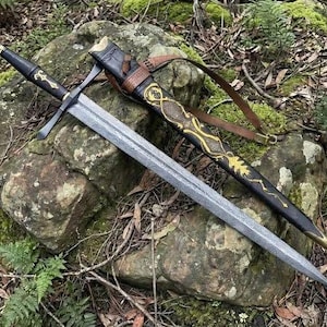 Può includere: Una spada in acciaio a doppio taglio con lama testurizzata, impugnatura rivestita in pelle nera con accenti dorati e guardia. Accanto si trova un fodero nero con motivi in filigrana dorata, punta in ottone e tracolla in pelle marrone.