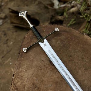 Könnte beinhalten: Ein Schwert aus silberfarbenem Metall mit einem schwarz umwickelten Ledergriff, einem kreuzförmigen Parier und einem Knauf mit dekorativen Details. Die Klinge zeigt ein eingraviertes Sternsymbol nahe dem Heft und Runenzeichen entlang der Hohlkehle.