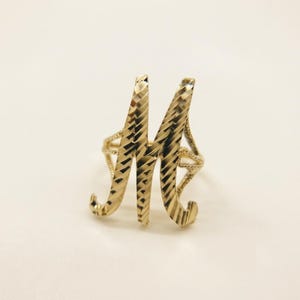 Może przedstawiać: Złoty pierścionek z dużą, stylizowaną literą M jako elementem centralnym. Powierzchnia litery M ma teksturowane wykończenie z diamentowym szlifem, które odbija światło, a obrączka pierścionka jest cienka i delikatna, łącząc się z literą.