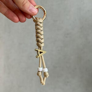 Könnte beinhalten: Ein beigefarbener Paracord-Schlüsselanhänger mit geflochtenem Design, einem goldfarbenen Metallring an der Oberseite, einem goldfarbenen Buchstaben-A-Anhänger in der Mitte und zwei weißen Perlen, die durch Knoten an den Enden befestigt sind.