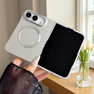 Puede incluir: Una funda para smartphone plegable de color plateado con un anillo de carga magnético circular en el panel posterior. La funda tiene un módulo de cámara elevado con dos lentes circulares y un pequeño flash, y una pantalla negra visible.