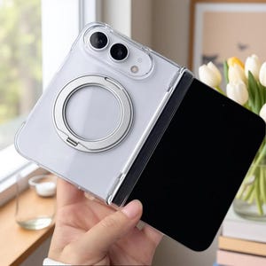May include: A clear, foldable smartphone case featuring a silver circular ring stand on the back panel. The case has precise cutouts for the dual camera lenses and flash, with a transparent design that reveals the phone's structure underneath.