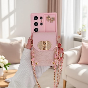 May include: A light pink smartphone case featuring a textured surface, a gold-toned metal turn-lock closure, and a decorative gold bow accent. The case includes a long, gold-toned metal chain strap interwoven with pink material for carrying.