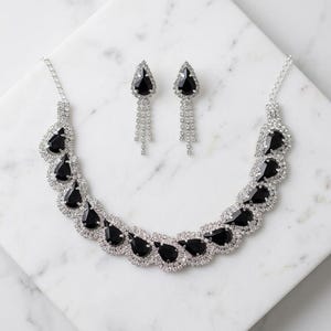 Könnte beinhalten: Ein Schmuckset bestehend aus einer Halskette und passenden Ohrringen. Die Halskette zeigt eine Reihe schwarzer tropfenförmiger Steine, die von klaren Kristallen umgeben sind. Die Ohrringe haben einen einzelnen schwarzen Tropfenstein.