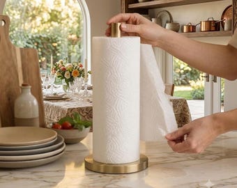May include: A vertical paper towel holder featuring a circular brushed gold metal base and a matching cylindrical top finial. The holder supports a standard roll of white embossed paper towels, which are being pulled by a hand.