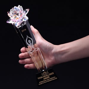 以下が含まれることがあります： ファセットカットされた蓮の花のトップ、ヨガのポーズのシルエットと2019年が刻印された垂直の柱、そしてYOGA EXCELLENCE AWARD INTERNATIONAL YOGA FEDERATIONという文字が記された黒い台座を持つ透明なクリスタルのトロフィー。