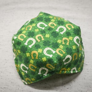 May include: Green fabric with a pattern of white horseshoes and four-leaf clovers. The fabric is folded into a triangular shape.