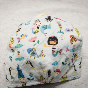 May include: A white fabric face mask with a colorful pattern of mermaids, fish, and sea creatures. The mask has a blue, pink, and green color scheme.