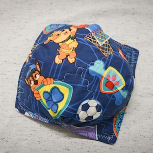 May include: A blue fabric face mask with a repeating pattern of cartoon dogs playing basketball. The mask features a yellow, orange, and red color scheme.