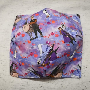 May include: Purple fabric face mask with a repeating pattern of characters from the animated movie Frozen II. The characters include Elsa, Anna, Kristoff, and Olaf. The background is a light purple with a white and pink swirl pattern.