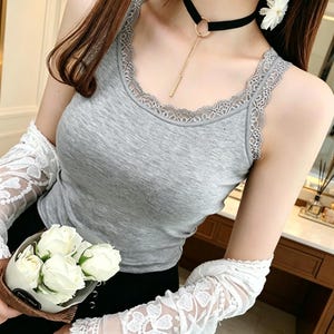 May include: A grey sleeveless tank top featuring a delicate lace trim along the scoop neckline and armholes. The garment is paired with white lace sleeves and a black choker necklace with a gold-toned chain pendant, alongside a small bouquet of white roses.