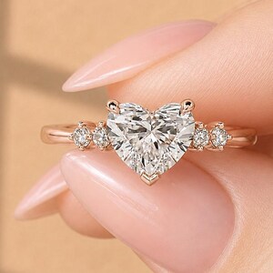 Puede incluir: Un anillo de oro rosa con un diamante central en forma de corazón montado en garras. La banda está adornada con dos pequeños diamantes redondos a cada lado de la piedra central, creando un diseño simétrico con un acabado metálico pulido.