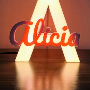 Può includere: Una grande lettera A illuminata con una trama a nido d'ape, che presenta il nome Alicia in una scrittura corsiva con una sfumatura dall'arancione al viola sovrapposta sul davanti. La luce emette un bagliore caldo, evidenziando la struttura.
