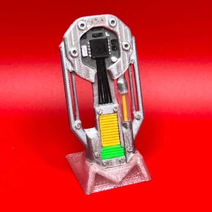 Pode incluir: Um acessório impresso em 3D de cor prateada com um componente central de placa de circuito preto com cablagem visível, uma secção amarela canelada, uma base retangular verde e um pequeno tubo transparente com uma haste amarela, numa base rosa.