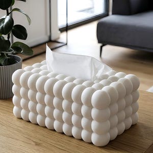 May include: A rectangular tissue box cover featuring a textured surface composed of numerous small, uniform white spheres arranged in a grid pattern. A single white paper tissue is pulled through the central opening at the top of the box.