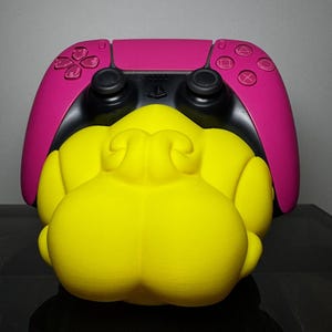 Può includere: Un controller per videogiochi rosa brillante appoggiato su un supporto giallo a tinta unita, arrotondato e con lobi multipli. Il controller presenta levette analogiche nere, un pannello centrale nero con logo PlayStation e tasti rosa.