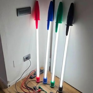 Könnte beinhalten: Vier hohe Stehlampen in Form von überdimensionalen Kugelschreibern. Jede Lampe hat einen klaren zylindrischen Körper mit hellem weißem Licht im Inneren, gekrönt von einer farbigen Kappe und Basis in Rot, Blau, Grün und Schwarz, verbunden mit Stromkabeln.