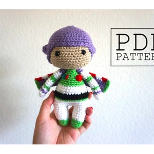 May include: Crocheted amigurumi toy of Buzz Lightyear from the Toy Story franchise. The toy is purple, green, white, and red. The toy is holding a red and white striped scarf. The text "PDF PATTERN" is in the upper right corner of the image.