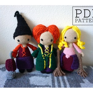 Puede incluir: Tres muñecas de crochet vestidas de brujas. La muñeca de la izquierda lleva un vestido morado, una camisa roja y un sombrero negro puntiagudo. La muñeca del medio lleva una túnica verde con detalles amarillos y una peluca roja. La muñeca de la derecha lleva un vestido rosa con una falda morada y una peluca amarilla. Las muñecas sostienen escobas y una aspiradora.