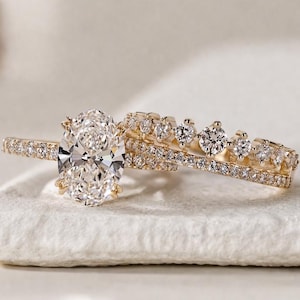 Könnte beinhalten: Ein Goldring-Set bestehend aus einem oval geschliffenen Mittelstein, der von kleineren Pavé-Diamanten am Ringband umgeben ist, sowie einem passenden Goldring mit einer Reihe abgestufter, rund geschliffener Diamanten am oberen Rand.