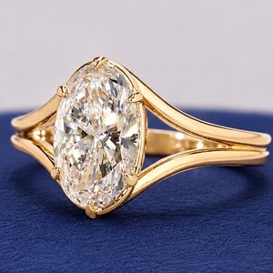 Könnte beinhalten: Ein goldener Ring mit einem großen, klaren, oval geschliffenen Edelstein in einer Krappenfassung. Die Ringschiene ist in zwei glatte, polierte goldene Arme geteilt, die an der Basis des zentralen Steins zusammenlaufen.