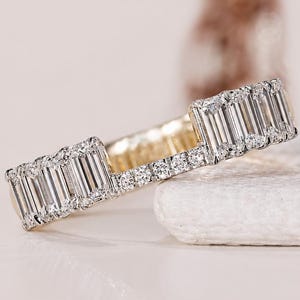 Könnte beinhalten: Ein goldener Ring mit einer Reihe von Diamanten im Baguetteschliff, akzentuiert durch kleinere runde Brillantschliff-Diamanten an den Rändern. Das Design weist eine deutliche Lücke zwischen zwei Abschnitten der Baguettesteine auf.