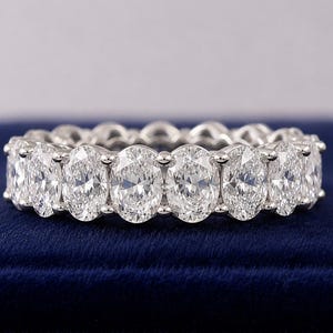 Peut inclure: Une alliance argentée ornée d'une rangée continue de pierres transparentes taille ovale, fixées par des griffes. La bague présente un alignement uniforme de gemmes à facettes qui captent la lumière, offrant un aspect brillant et réfléchissant.