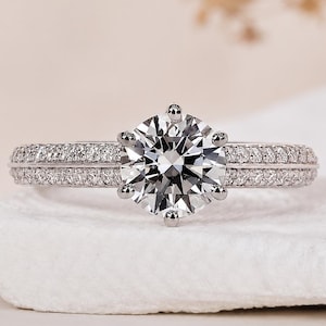 Könnte beinhalten: Ein silberfarbener Ring mit einem großen, runden Brillantschliff-Stein, der von sechs Krappen gehalten wird. Die Ringschiene ist in zwei parallele Reihen unterteilt, die jeweils mit kleinen, klaren, runden Steinen besetzt sind.