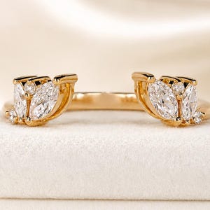 Könnte beinhalten: Ein offener Goldring mit zwei symmetrischen Clustern aus marquise- und rundgeschliffenen klaren Steinen. Das Band besteht aus poliertem Gold und die beiden Enden sind mit blattförmigen Steinanordnungen verziert, die sich gegenüberstehen.