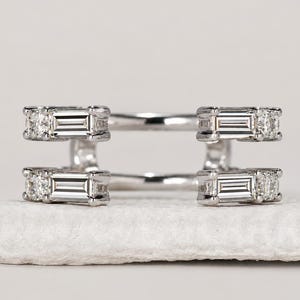May include: A silver-toned ring guard featuring two parallel bands connected by vertical bars. Each end of the bars is set with a rectangular baguette-cut stone flanked by a small round brilliant-cut stone, creating a symmetrical open design.