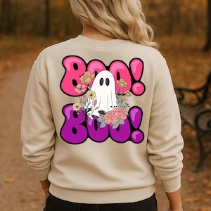 Könnte beinhalten: Ein beigefarbenes Sweatshirt mit Rundhalsausschnitt und einem Grafikdruck auf der Rückseite. Das Design zeigt ein weißes Gespenst, umgeben von bunten Blumen, sowie den Schriftzug BOO! in leuchtend pinken und violetten Buchstaben mit schwarzem Rand.