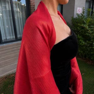 May include: A bright red, textured shawl draped over a black strapless dress. The fabric of the shawl features a fine, crinkled, pleated pattern with a subtle sheen. A thin gold chain necklace with a small pendant rests against the collarbone.