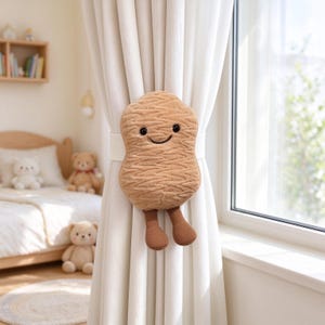 May include: A tan, plush, nugget-shaped curtain tieback featuring a smiling face with black embroidered eyes and a mouth. The soft, textured character has two dangling, ribbed brown legs and is attached to a white elastic band holding a curtain.