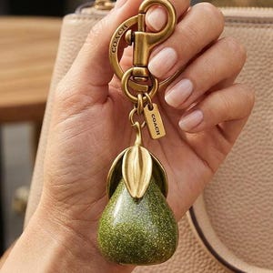 May include: A pear-shaped keyring featuring a glittery green body with a metallic gold-tone stem and leaves. The charm is attached to a brass-toned lobster clasp and a small rectangular tag engraved with the word COACH in capital letters.