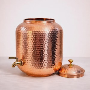 May include: A large, cylindrical copper beverage dispenser featuring a distinct hammered texture across its exterior surface. The vessel includes a brass-colored metal spigot attached to the front and a matching circular lid with a small knob handle.
