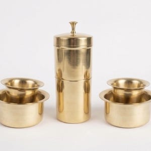 May include: A polished brass coffee filter set consisting of a tall cylindrical brewing vessel with a lid and two matching wide-rimmed cups. The metallic gold-toned kitchenware features a smooth finish and a classic multi-part construction.