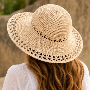 May include: A beige straw sun hat featuring a rounded crown and a wide brim with an intricate, open-weave crochet pattern along the edge. The hat is constructed from a woven material with a consistent light tan color throughout the design.
