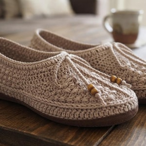 May include: A pair of beige crochet slippers featuring a textured stitch pattern, a rounded toe, and a decorative drawstring closure at the front adorned with small, smooth wooden beads. The slippers have a solid, flat brown sole for support.