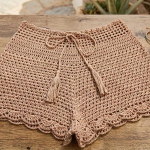 May include: Beige crochet shorts featuring an open-knit pattern, a drawstring waistband with two dangling tassels, and a scalloped hemline. The shorts are displayed alongside two thin, circular, metallic-toned bracelets on a wooden surface.