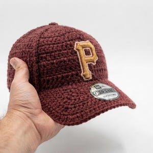 以下が含まれることがあります： ゴールドとホワイトの刺繍で文字Pが施された、マルーンカラーのかぎ針編みベースボールキャップ。キャップのつばには、スタイルとブランドを示すNew Era 9FORTYというテキストが書かれた、円形のシルバーとブラックのステッカーが貼られています。