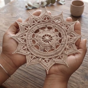 May include: A circular beige crochet doily featuring an intricate star-shaped pattern with a central wheel design, radiating outward into pointed edges. The piece is crafted with fine thread, showcasing detailed stitching and a symmetrical geometric layout.