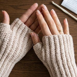 May include: A pair of beige fingerless gloves featuring a vertical ribbed knit pattern. The gloves cover the wrist and palm area while leaving the fingers exposed. A light-colored crochet hook and a small paper pattern guide are also visible.