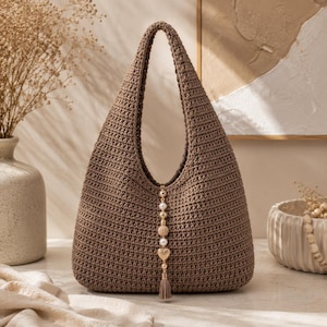 Peut inclure: Un sac hobo en crochet marron doté d'une anse intégrée unique et d'une breloque décorative verticale centrale. La breloque se compose d'une série de perles dorées, de fausses perles blanches et d'un petit pendentif cœur doré avec un pompon.