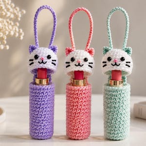 May include: Three crochet lipstick holders featuring cat-shaped caps with black eyes, pink noses, and whiskers. Each holder has a matching loop handle and contains a lipstick tube. The holders are colored in light purple, pink, and mint green.
