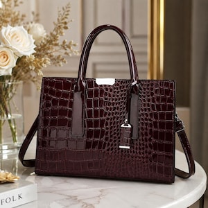 May include: A dark burgundy handbag with a glossy finish and a textured crocodile skin pattern. The bag features two top handles, a rectangular silhouette, and a small silver-toned metal accent plate at the centre top of the front panel.