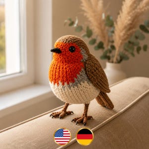 May include: A crocheted robin bird figurine featuring a bright orange chest, a brown head and wings, a cream-colored belly, and a small black beak. The bird has dark eyes and brown yarn feet with visible claws, standing upright on a textured surface.
