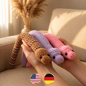 May include: Three plush crochet snakes in brown, lavender, and pink. The brown snake features a repeating pattern of white and dark brown stripes. Each snake has small black bead eyes and a soft, textured yarn construction with a rounded head.