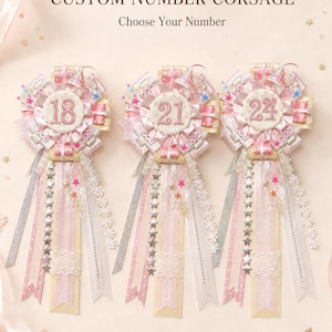 May include: Three custom number corsages featuring the numbers 18, 21, and 24. Each round badge is decorated with pink and white pleated ribbons, small party hat accents, and long hanging streamers with star patterns and lace trim.